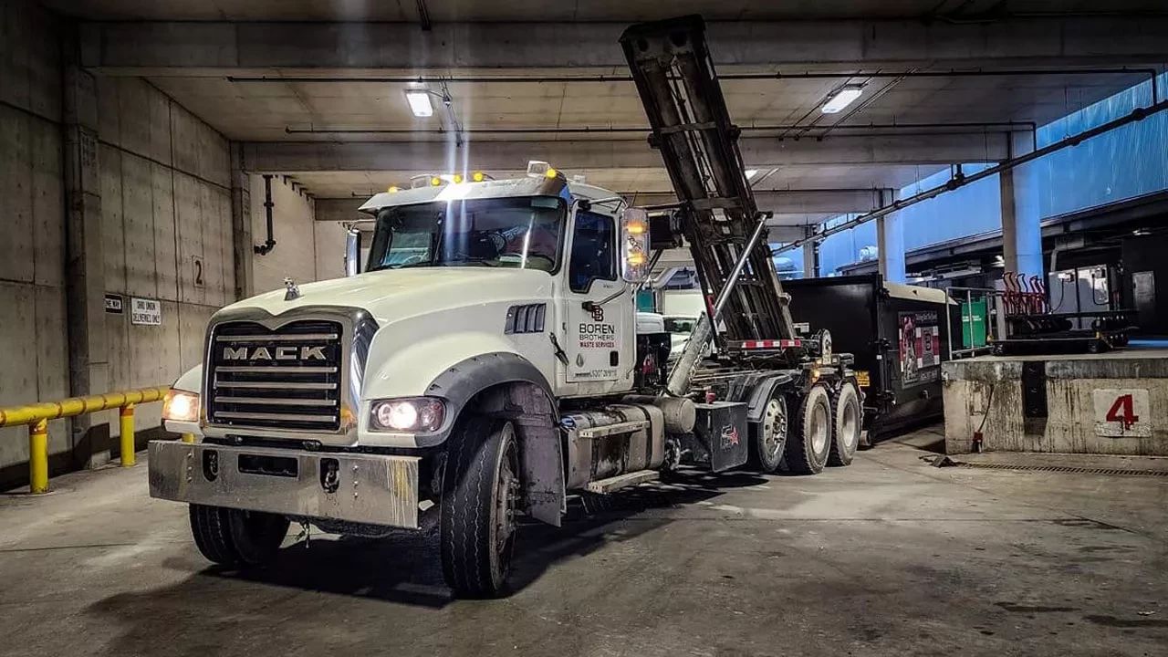 A Boren Brothers Waste Services Mack truck with its container lift raised is parked inside an industrial facility, ready for a dumpster pickup or drop-off.