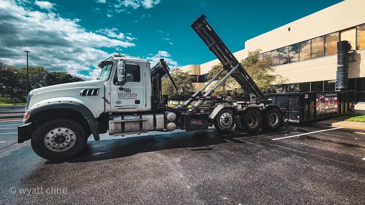 A Boren Brothers Waste Services truck is parked with its container lift raised, ready to load or unload a dumpster at a commercial site on a clear, sunny day.