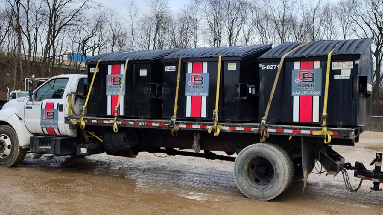 A Boren Brothers truck is parked on a muddy site, carrying multiple large black dumpsters securely strapped onto its flatbed, ready for delivery or pickup.